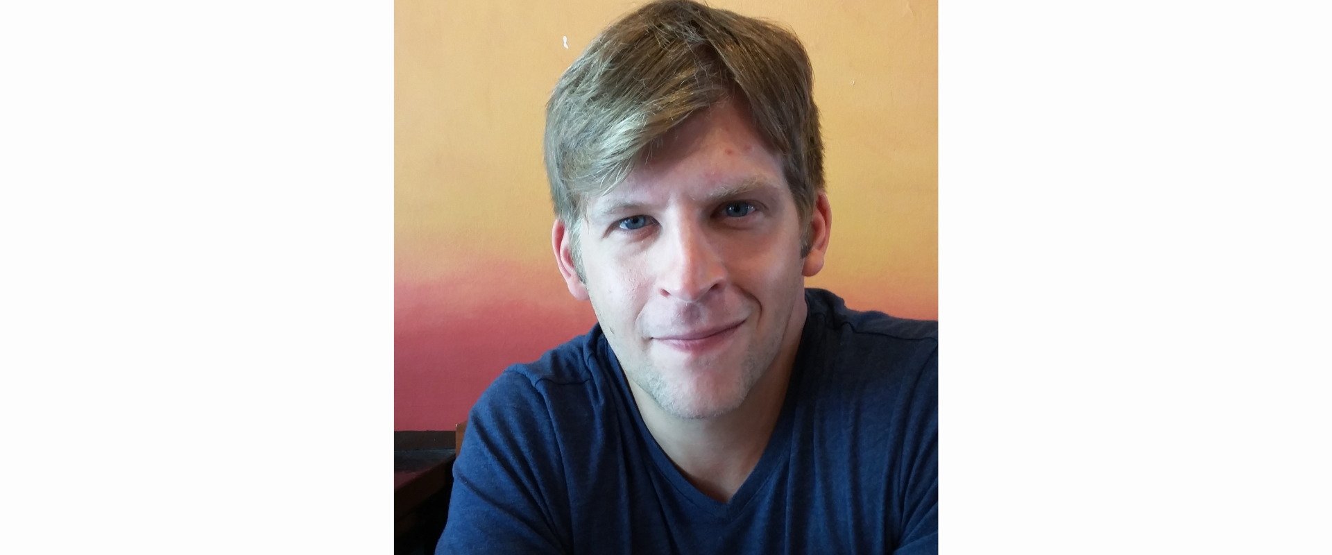 A person seated indoors wearing a dark blue short-sleeved shirt, positioned in front of a warm-toned wall that fades from orange to red.