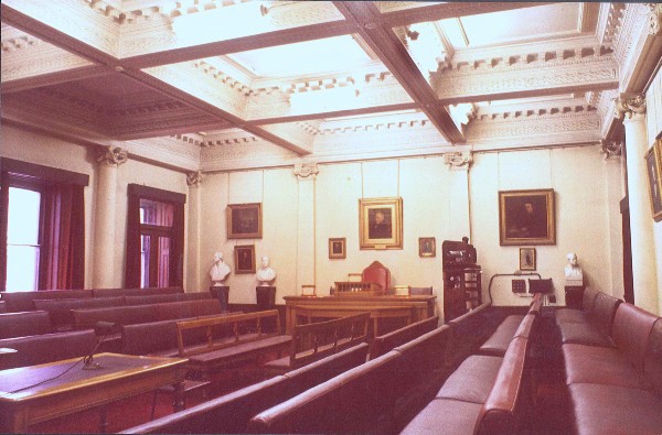 an old fashioned meeting room with dark burgundy sofas either side, tall sash windows and portraits on the walls