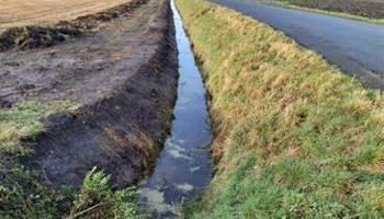 "A narrow water channel bordered by patches of grass and mud, featuring clear water reflecting sunlight, set against a dry landscape."