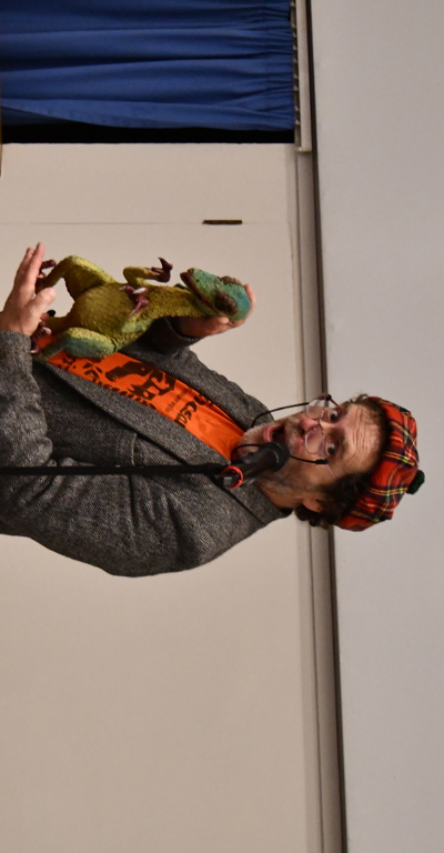A man wearing glasses, a tartan hat and a grey jacket holding a green dinosaur puppet