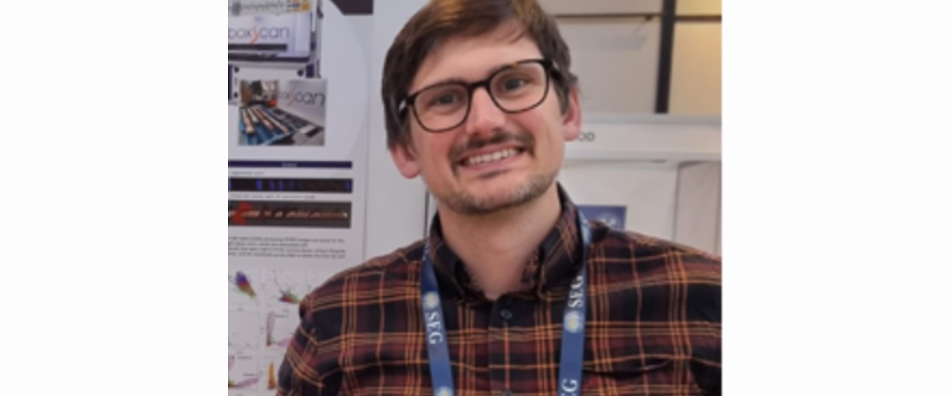 Person wearing glasses and a plaid shirt with a conference lanyard, standing in front of technical posters displayed on a board.