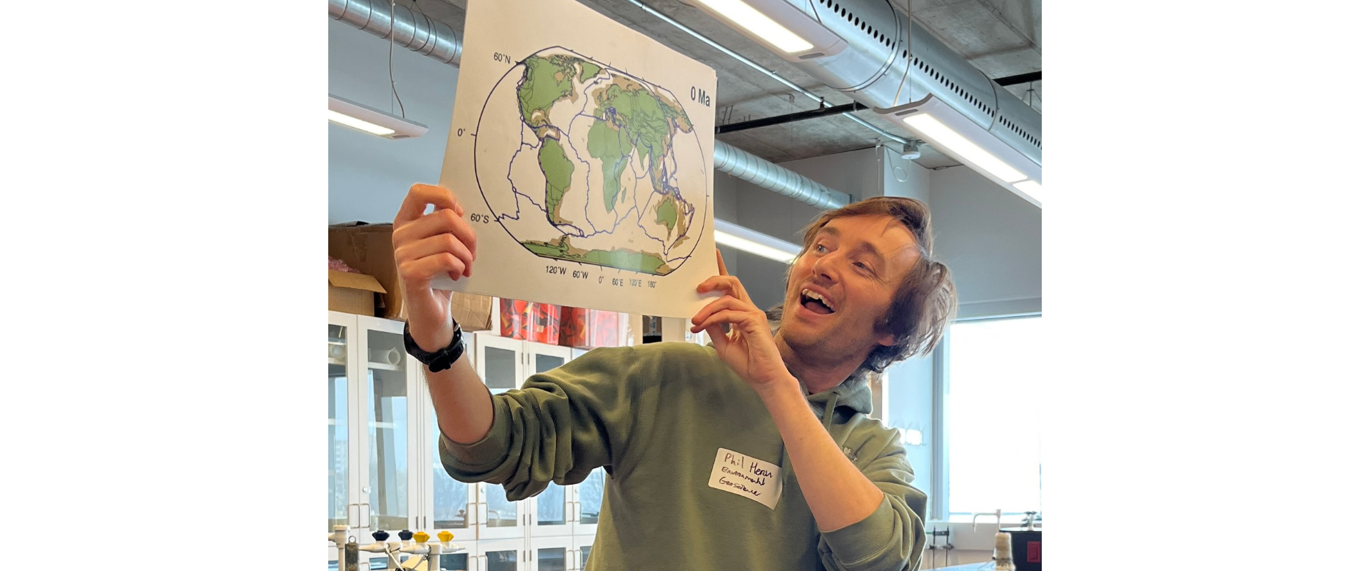 erson standing in a laboratory classroom holds up a printed map of the world showing tectonic plate boundaries and continental positions. The laboratory contains benches, sinks, cabinets, and overhead ductwork. A handwritten name badge is visible on the person's sweatshirt. Bright indoor lighting illuminates the scene.