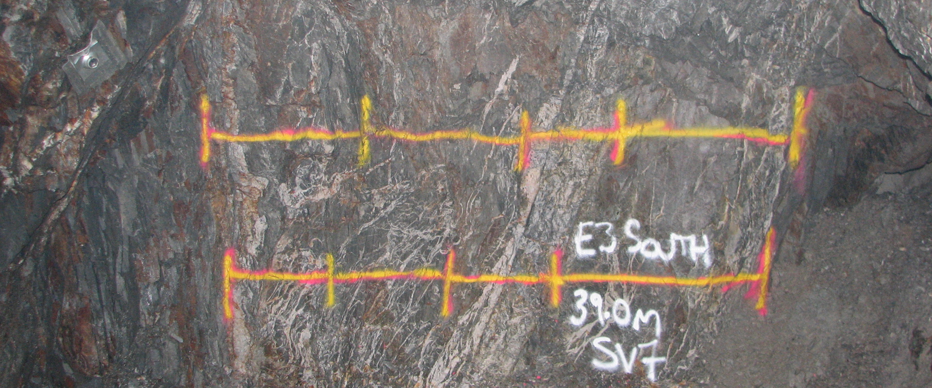 Underground rock wall with dark veined stone marked by bright yellow and pink painted horizontal lines with tick marks, indicating measured sections. White text reads “E3 South 39.0m SV7,” with loose debris and a cable visible at the base.