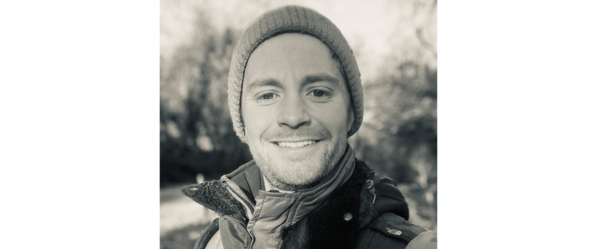 Black and white up portrait of a person outdoors, wearing a knitted beanie and layered jacket with visible backpack strap. The person faces the camera. Background shows blurred trees and a path, creating shallow depth of field and soft natural lighting throughout the scene with gentle contrast and even tones.