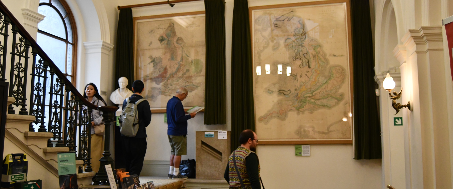 a stairway with two huge maps on the wall behind with a woman walking up the stairs and three men looking at the maps