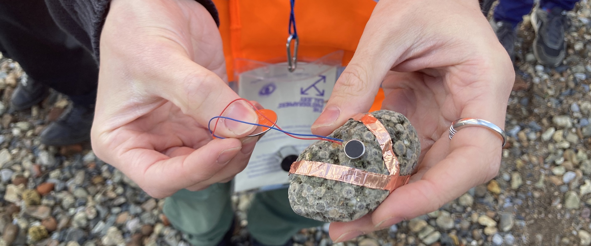 Hands holding a rock with copper wrapped in copper threads, connected to two wires and a coin