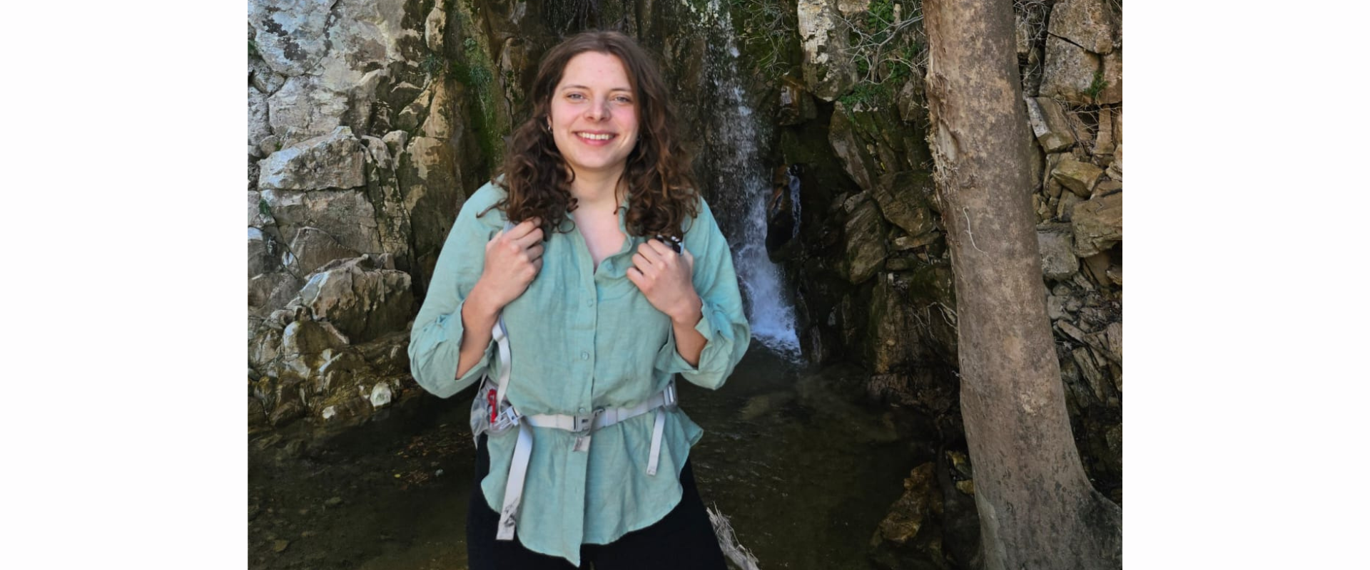Person wearing a light green long-sleeve shirt, black shorts, and a small backpack stands on rocks in a narrow, rocky area with a shallow pool and a small waterfall behind them; a tree trunk is visible on the right, and the surrounding rock face is covered with patches of moss.