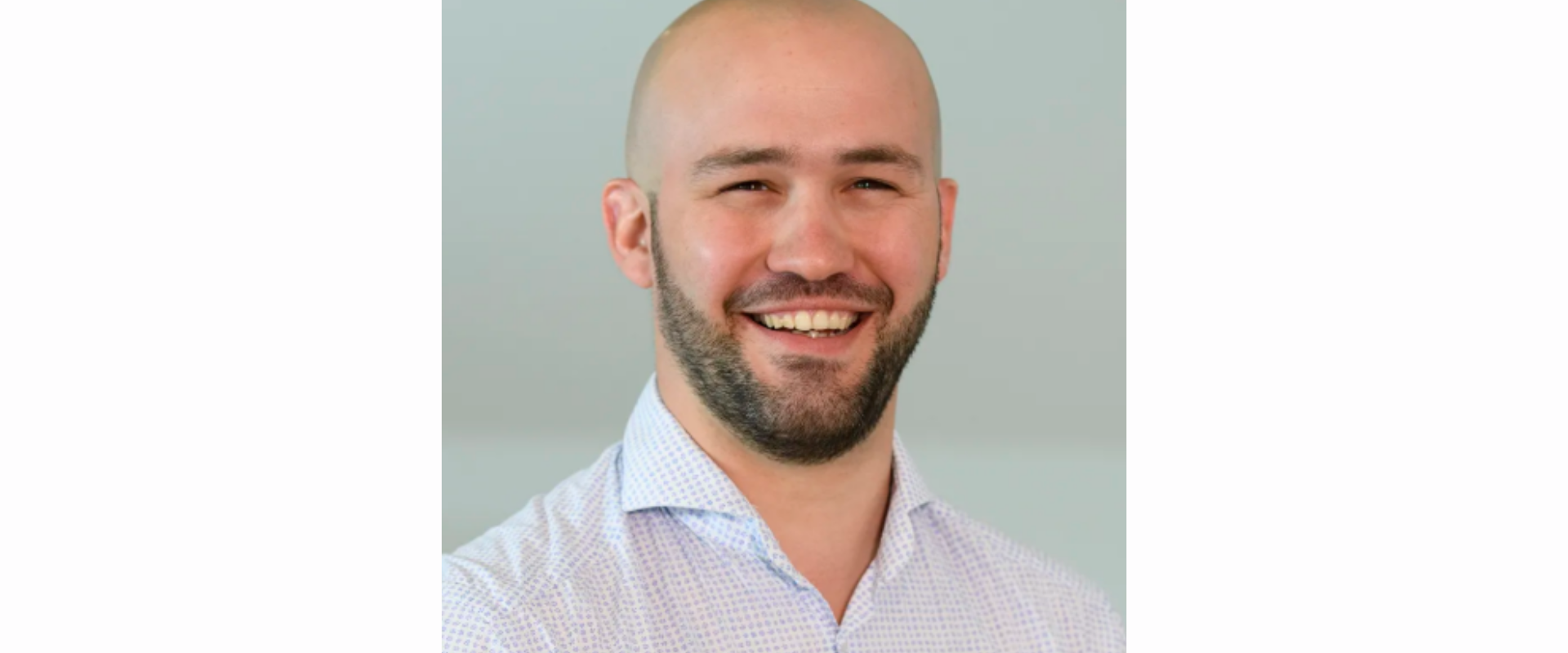 A person wearing a light-coloured, patterned collared shirt, visible from the chest upward, standing against a plain indoor background.