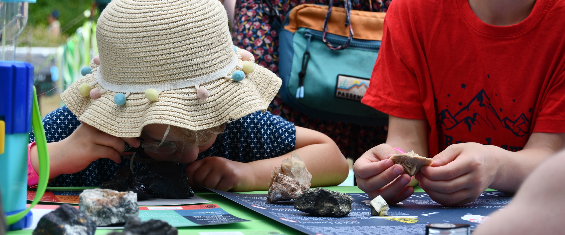 A kid with a hat looks at a rock through a magnifying glass, while another kid handles a rock on a table