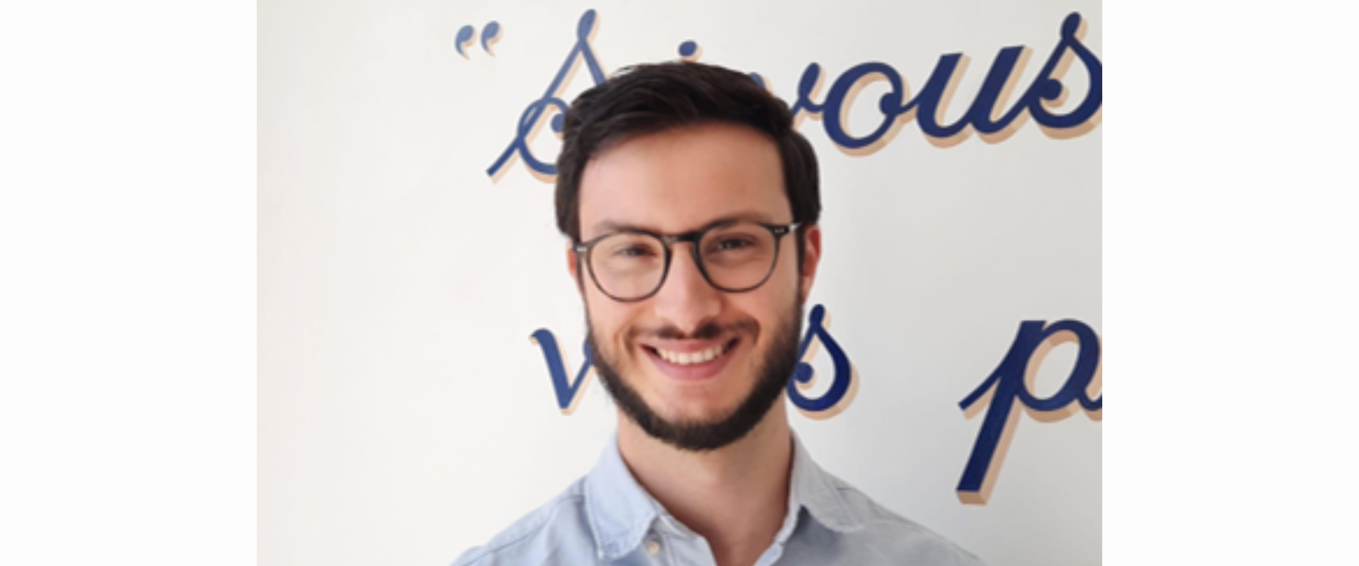 A person wearing a light button‑down shirt standing in front of a white wall with stylised blue script lettering in the background.
