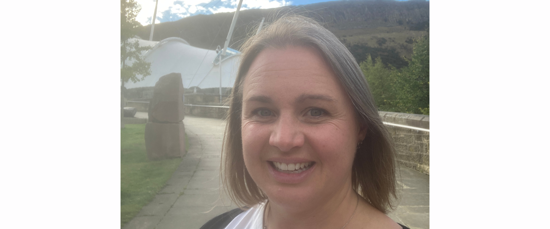 A person standing outdoors on a paved pathway with grassy areas and stone structures nearby. Behind them is a large white tensile canopy structure and a hillside under a bright blue sky with scattered clouds. The person is wearing a white top with a dark outer layer.