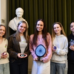 Five young women stand smiling with small geological samples in their hands. Behind them is a statue and a green velvet curtain, creating an academic atmosphere.