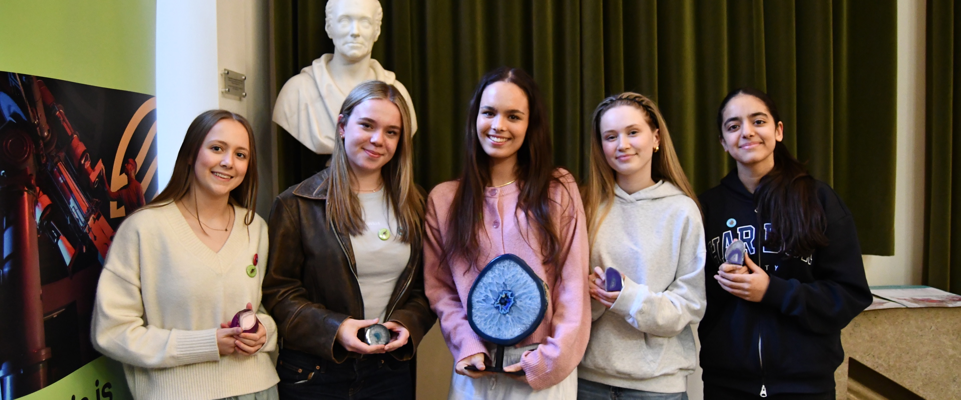 Five young women stand smiling with small geological samples in their hands. Behind them is a statue and a green velvet curtain, creating an academic atmosphere.