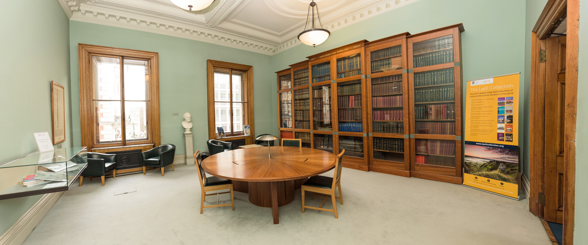 A reading room with a round wooden table, chairs, a glass display case, and tall bookshelves lining a wall, featuring a standing display sign for The Lyell Collection.
