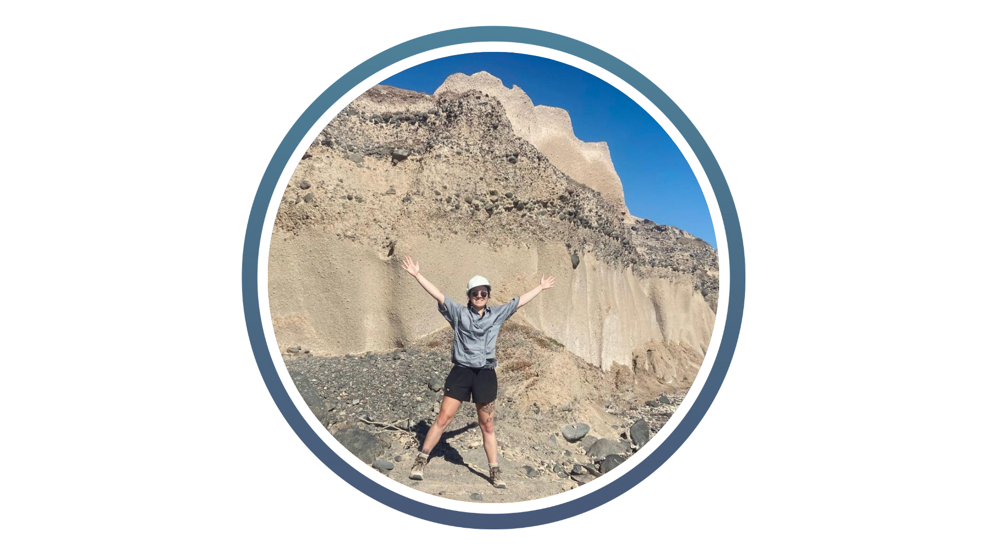 A person wearing a white helmet and casual outdoor clothing stands joyfully with arms outstretched in front of a large rock formation under a clear blue sky.