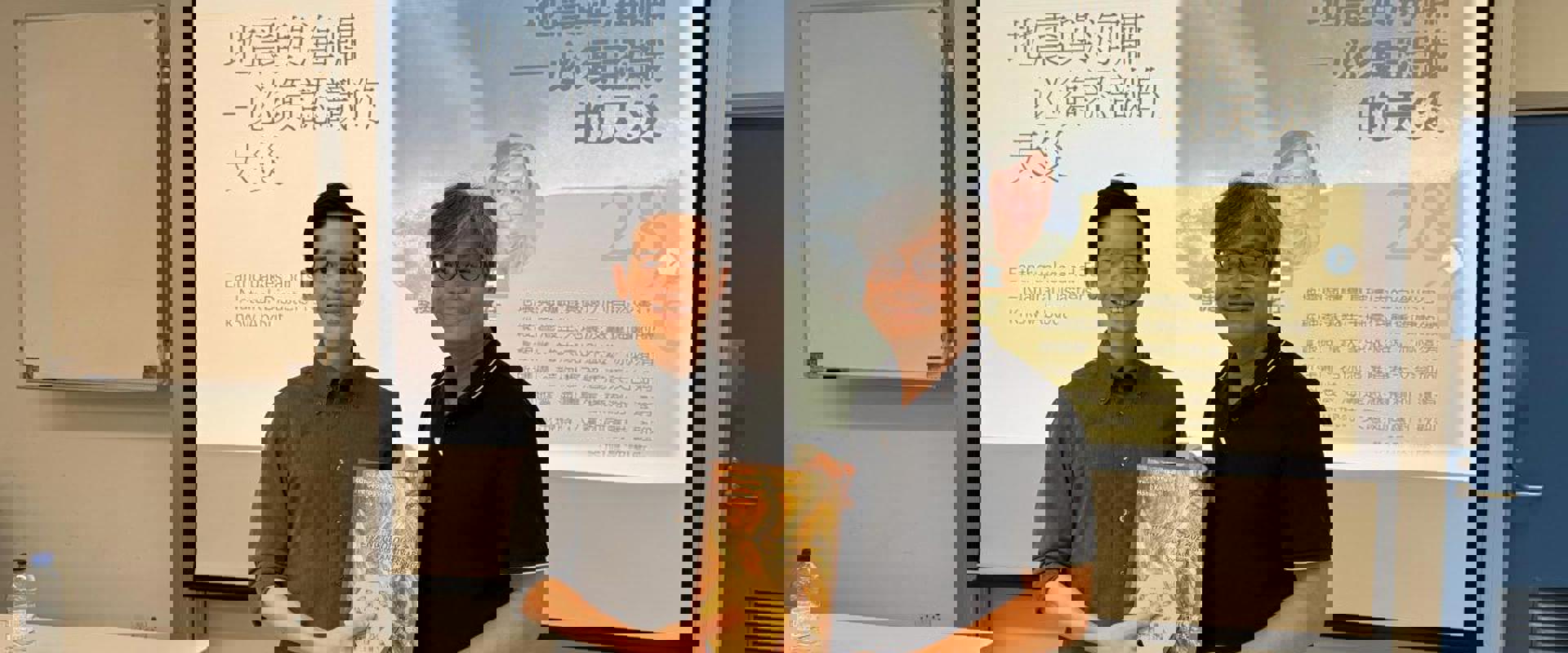 Two individuals stand in front of a presentation about natural disasters, one holding a book. A water bottle and table are visible.