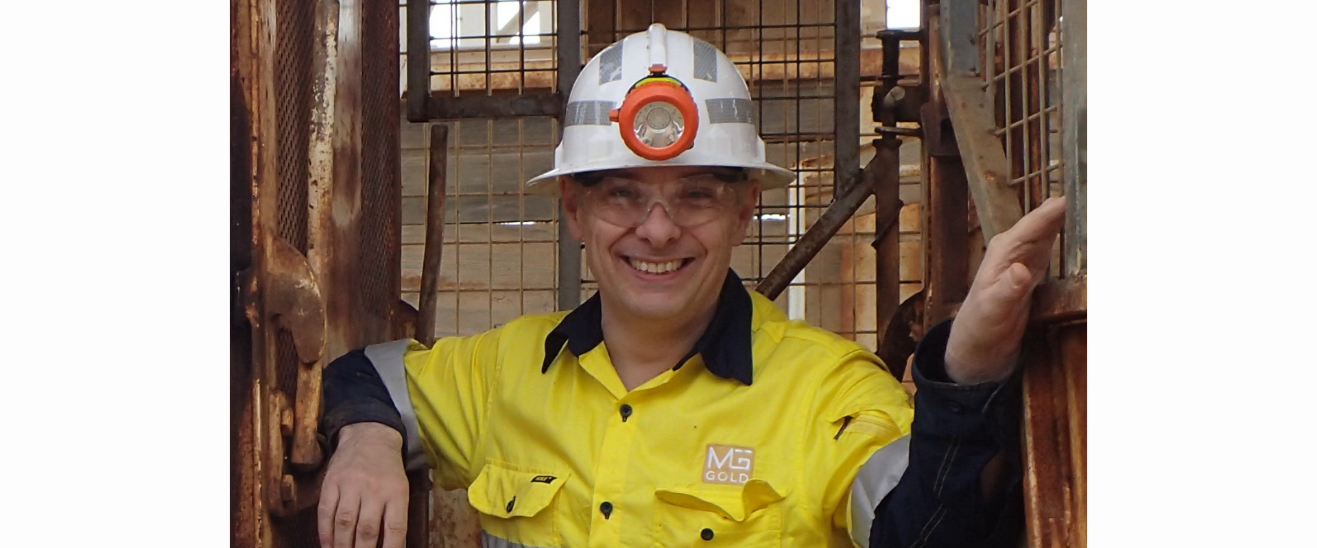 Alt text: Person wearing a white hard hat with headlamp, clear safety glasses, and a high‑visibility yellow work shirt, standing within a metal-framed underground mining environment.