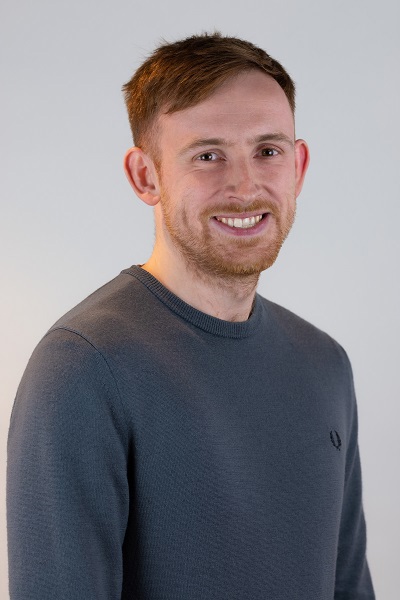 A man with short light brown hair and a trimmed beard is smiling at the camera, wearing a grey crew-neck sweater against a plain light background.