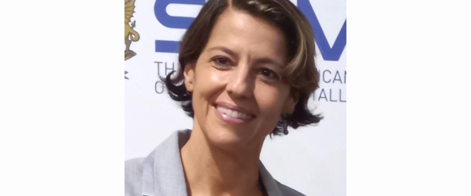 The image shows a close-up portrait of a single person positioned slightly off-center to the right. The setting appears to be a professional or corporate environment, likely at an event or press backdrop, indicated by a printed banner behind the person.