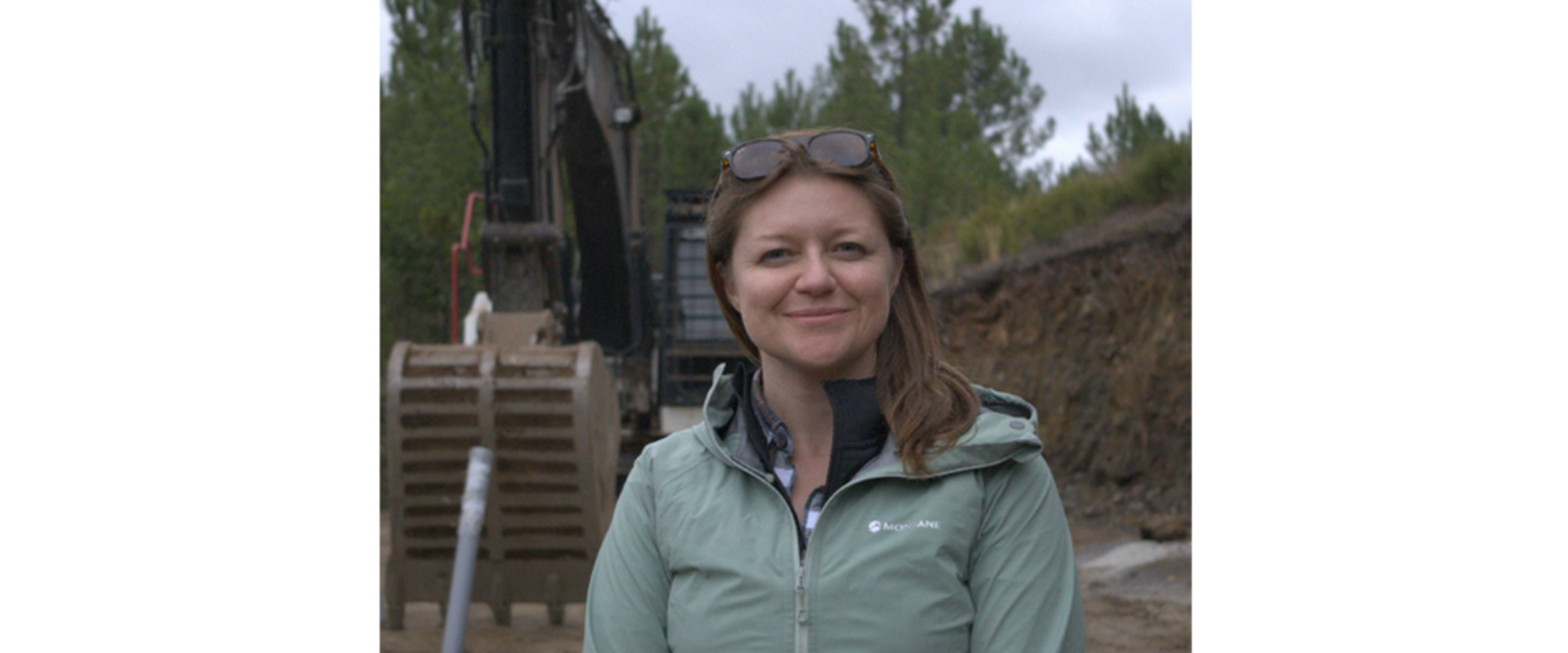 Person standing outdoors at an excavation site, wearing a light green zip-up jacket with sunglasses resting on the top of the head. An excavator bucket and digging equipment are visible behind the person, with a trench cut into the ground on the right and pine trees in the background under an overcast sky. Visible text on the jacket reads “Montane.”