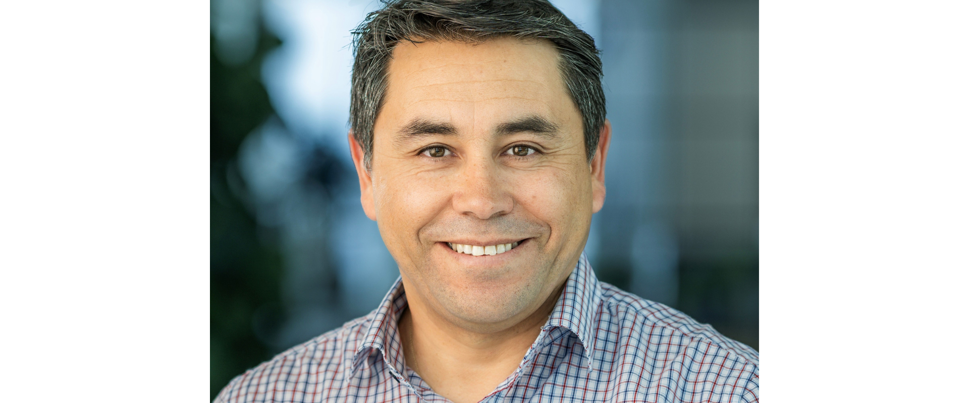 Close-up headshot of a person with short dark hair, wearing a checkered button-down shirt, facing the camera against a softly blurred indoor background.