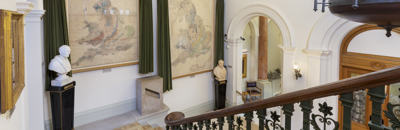 The staircase of the Geological Society of London, leading down stairs to the Society's entrance hall, with two large geological maps on the walls, and busts of George Greenough and William Smith