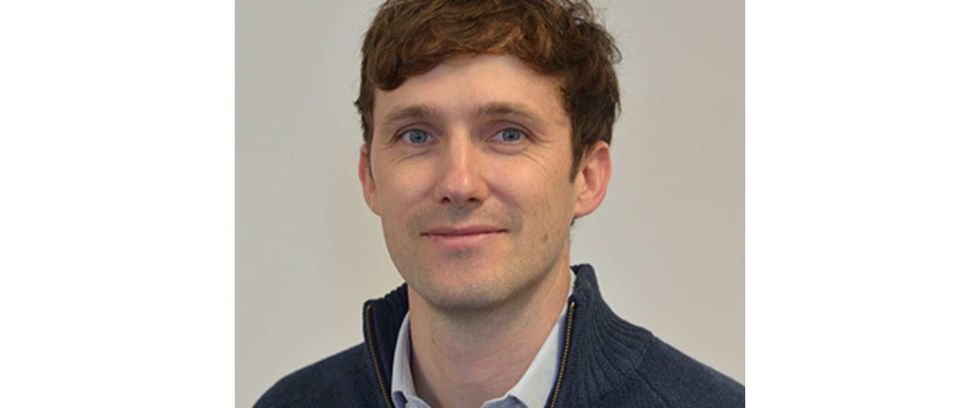 Head-and-shoulders portrait of a person facing the camera against a plain light background. The person has short, slightly wavy hair and wears a light collared shirt under a dark zip-up sweater. Lighting is soft and even.