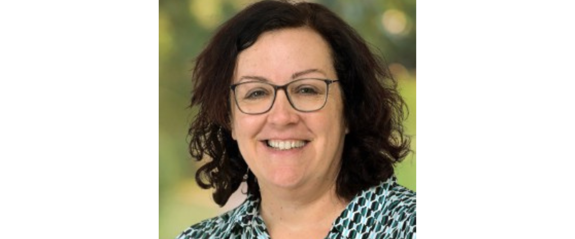 Head-and-shoulders professional portrait of a person with dark, shoulder-length curly hair and rectangular glasses, wearing a patterned blue, black, and white collared blouse. The person is facing the camera against a softly blurred green outdoor background with natural lighting.