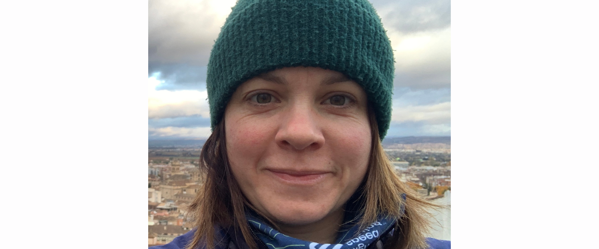 Close-up outdoor selfie of a person wearing a dark green knitted beanie with a pom‑pom and a blue jacket. A patterned neck scarf is visible at the collar. The person’s face fills most of the frame, with a cloudy sky and a cityscape of buildings and hills in the background, suggesting an elevated viewpoint.