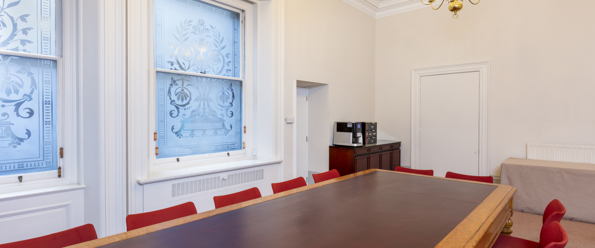 A spacious meeting room featuring a long wooden table, red chairs, decorative windows, and a coffee station in a bright setting.