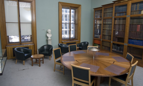 room with sash windows and a round table with seating and leather bucket armchairs and a small coffee table. a book case lines one wall behind