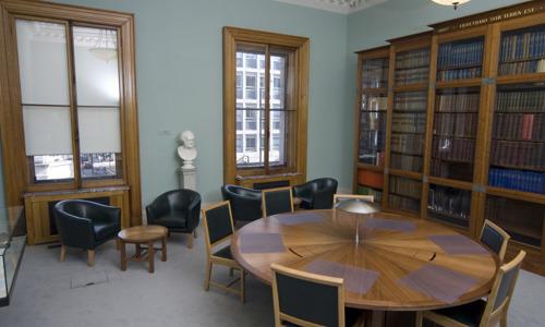 room with sash windows and a round table with seating and leather bucket armchairs and a small coffee table. a book case lines one wall behind