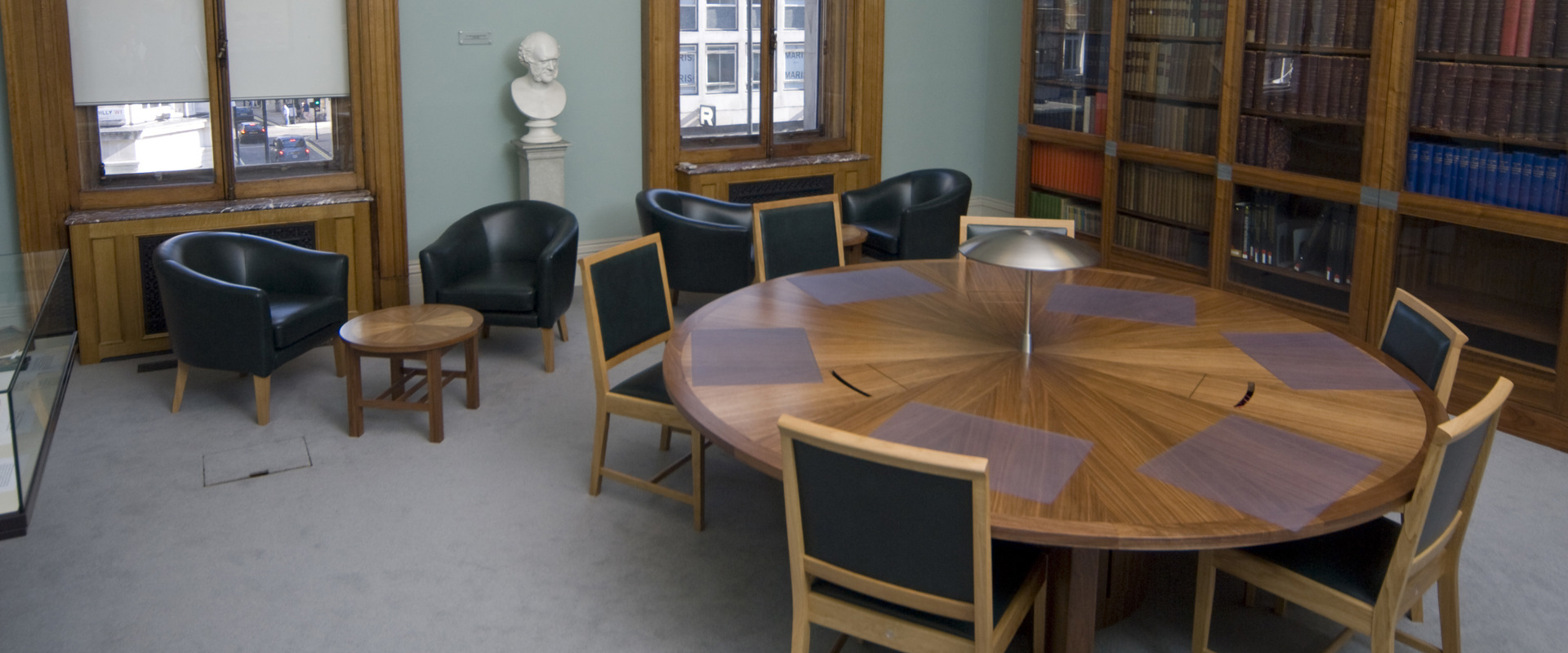 room with sash windows and a round table with seating and leather bucket armchairs and a small coffee table. a book case lines one wall behind