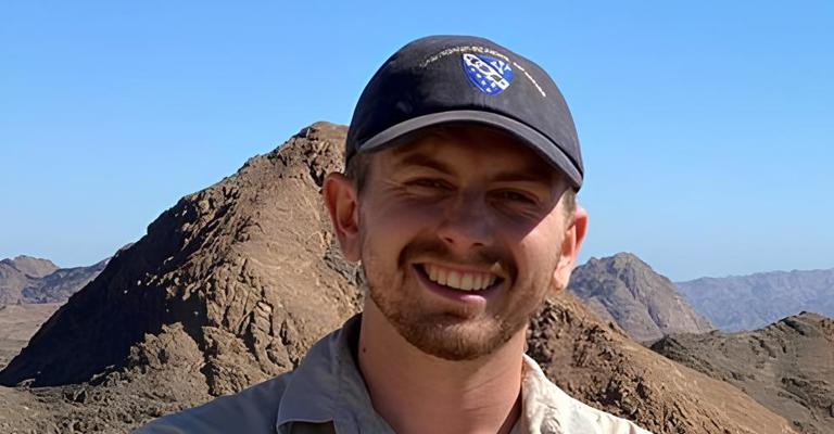 young white man with black cap standing in front of a mountain background with blue sky