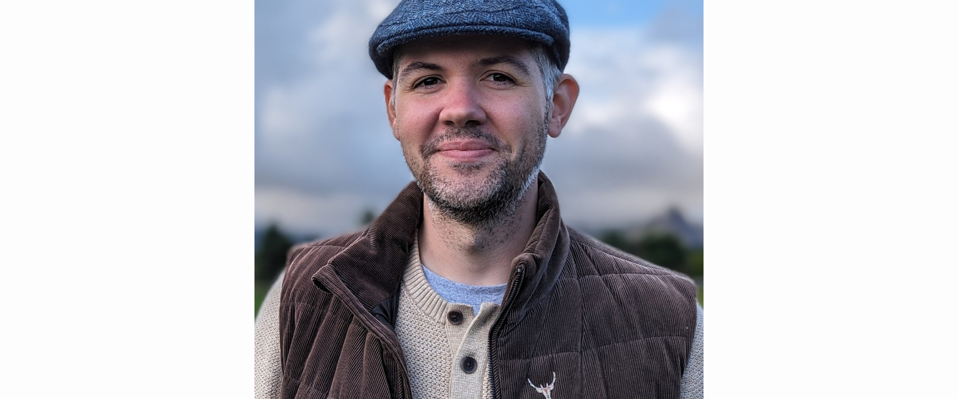 A person standing outdoors wearing a flat cap, a brown quilted vest over a light-coloured henley-style shirt, with a cloudy sky and soft landscape visible in the background.
